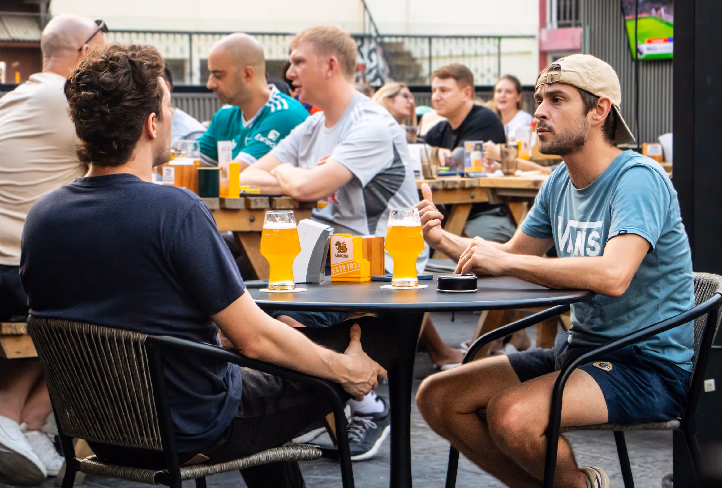Two men sit at a table with pints of beer, talking at an outdoor bar with other patrons nearby.