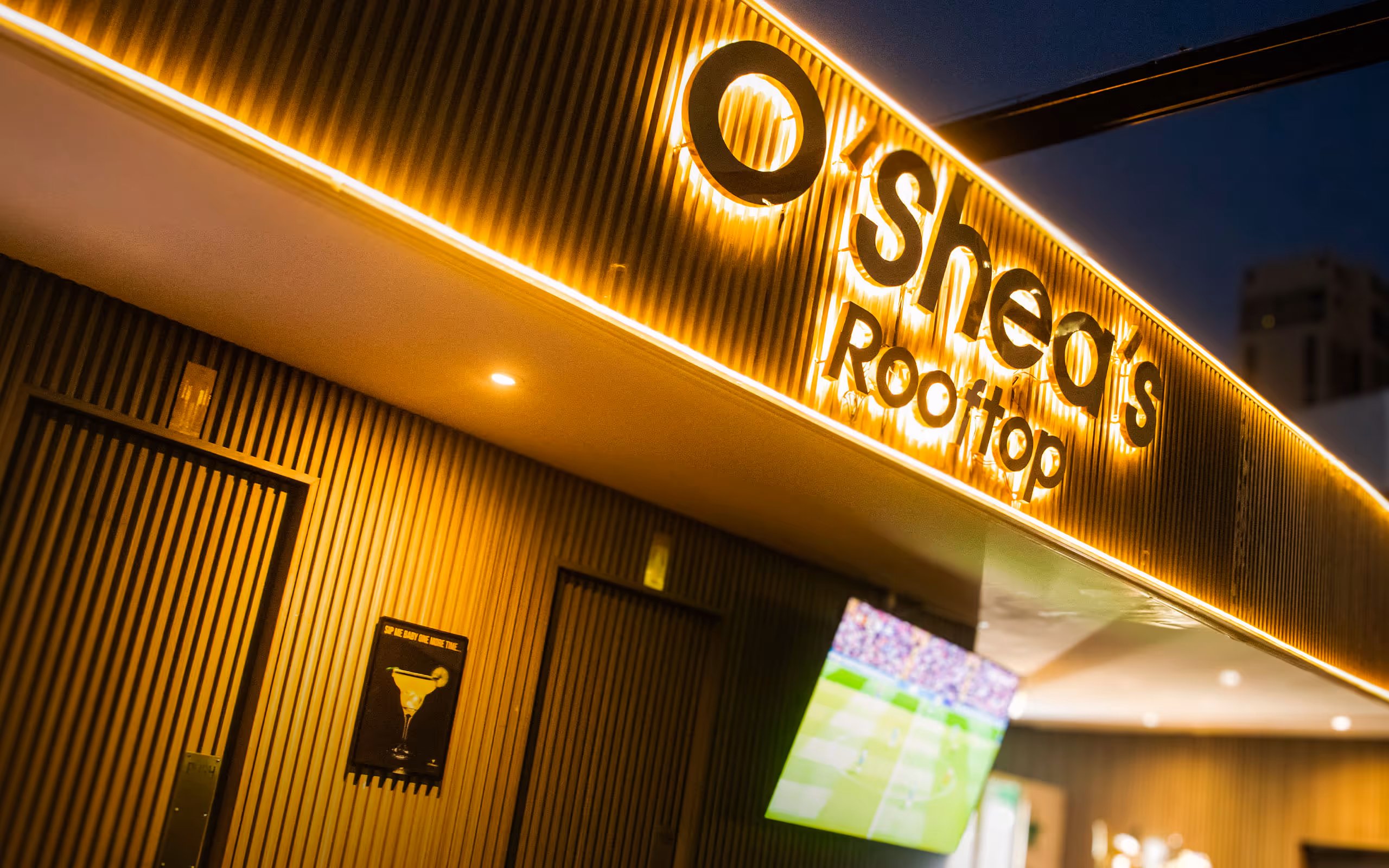 O'Shea's Rooftop sign lit with golden lights above a bar area showing a TV screen.