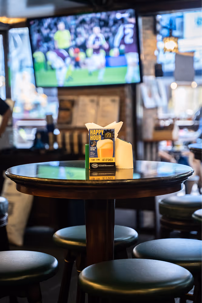 Round wooden table with green stools and a happy hour sign in a dimly lit bar with a TV in the background.