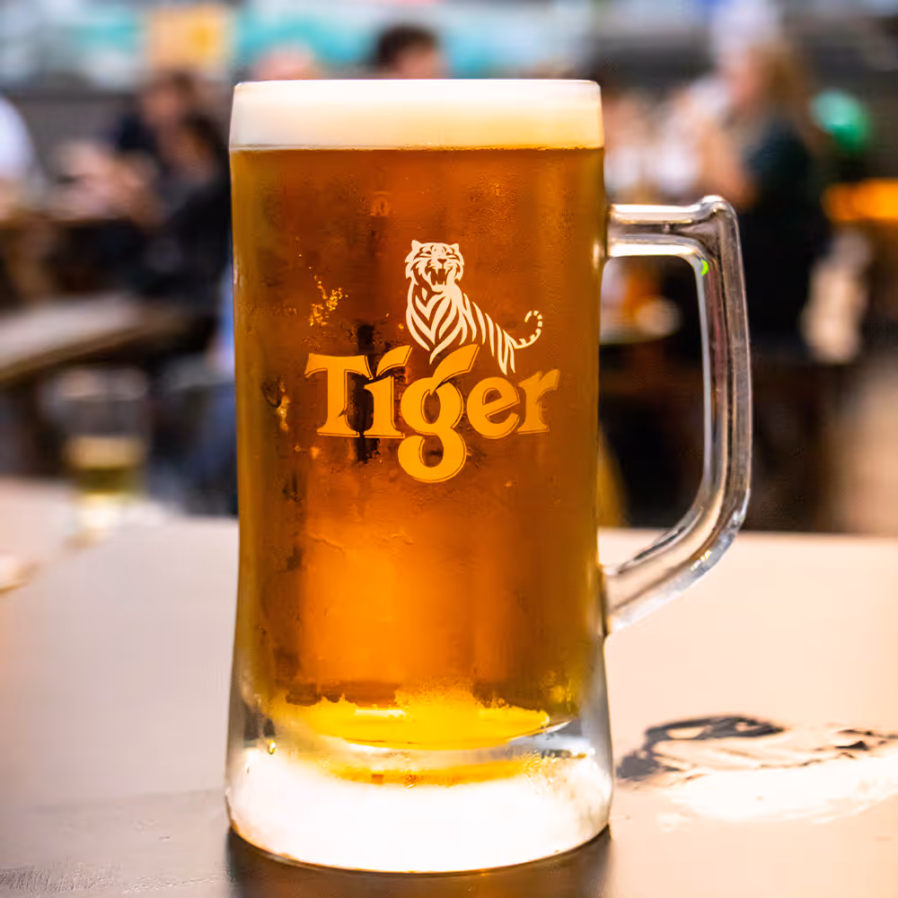 Frosted glass mug of Tiger beer with foam on top on a table in a bar setting.