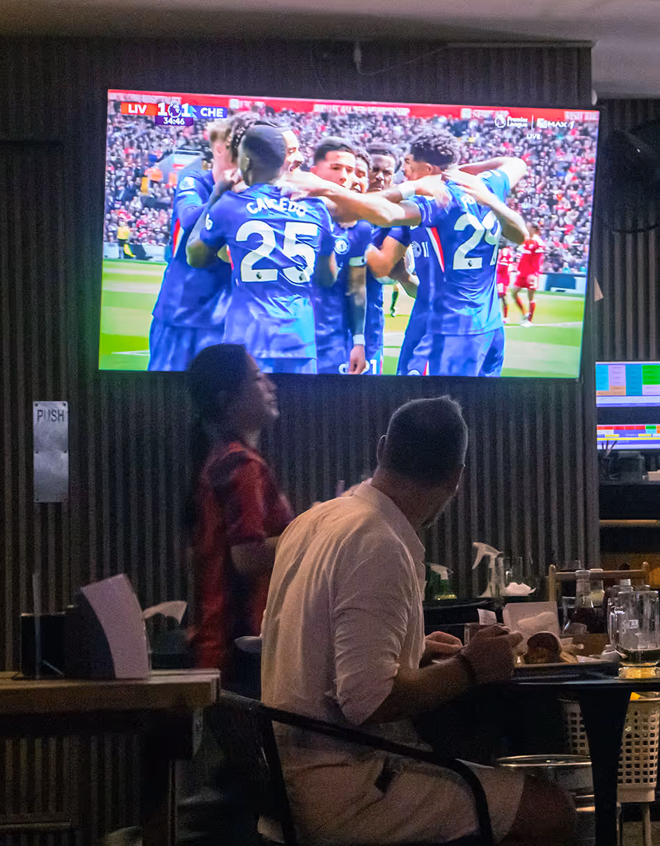 Bar patrons watch a soccer match on a TV showing Chelsea players celebrating a goal.