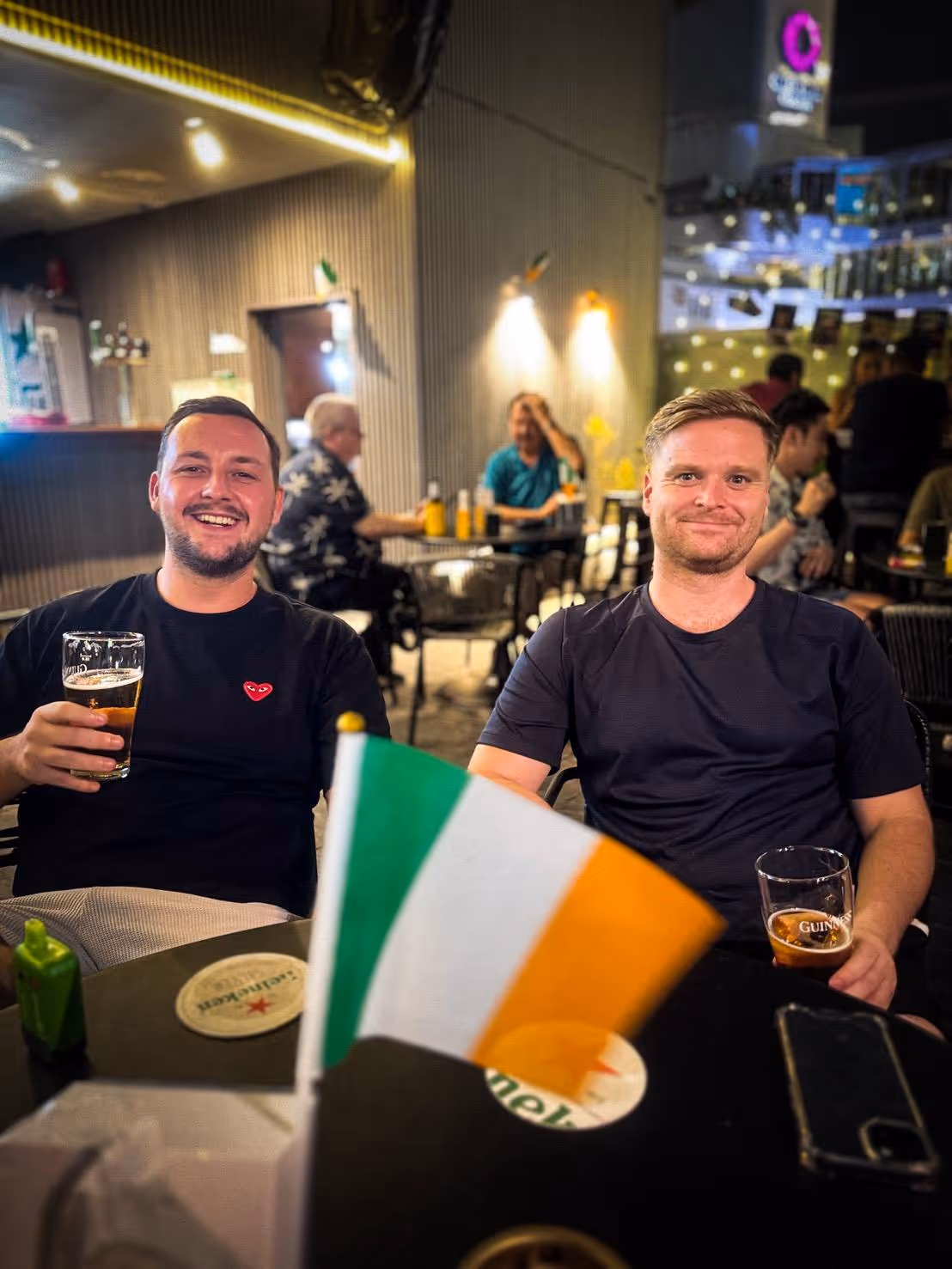 Two men sitting at a table in a pub holding beers with a small Irish flag in the foreground.
