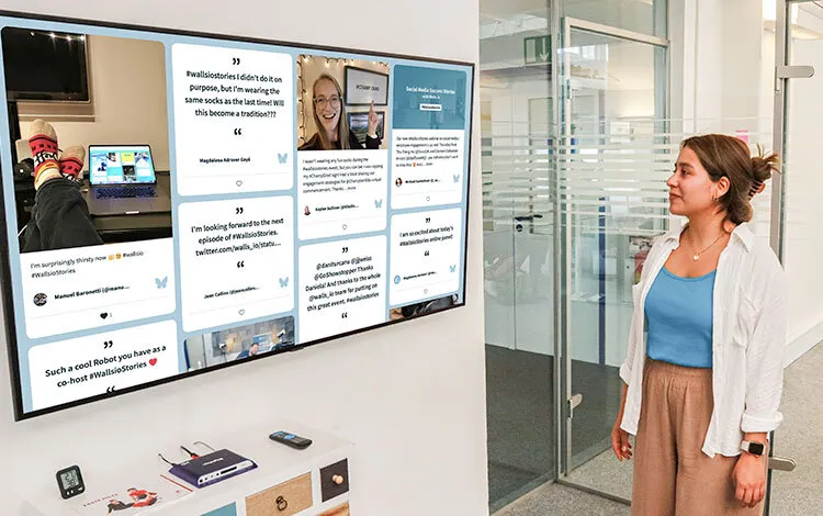 A woman observing a large digital screen displaying a bluesky feed with user-generated content in an office setting.
