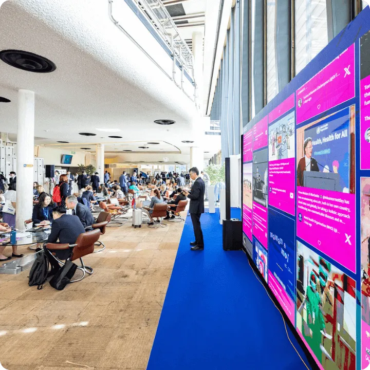 an event social wall displayed on a big screen. A man walking past it and other attendees in the background
