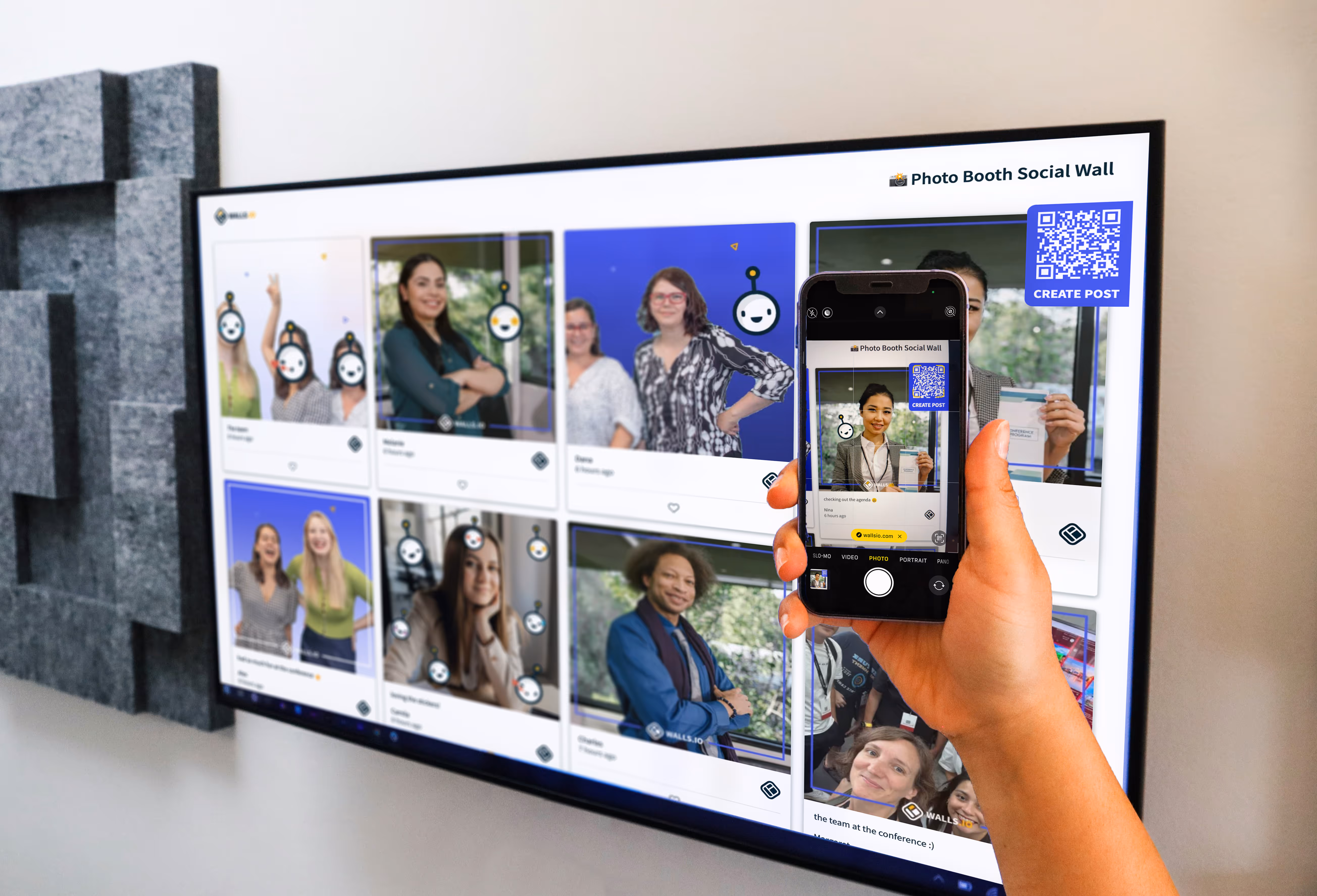 A large screen displays the Walls.io Photo Booth Social Wall, showing attendee photos from an event. In the foreground, a person holds up a smartphone capturing one of the displayed posts with the same layout and QR code visible.