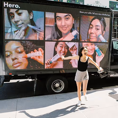 An outdoor digital signage on a truck in the times square displaying user generated content from the hero cosmetics company campaign. a customer who participated is very happy to see her photo there and is pointing happy towards the screen