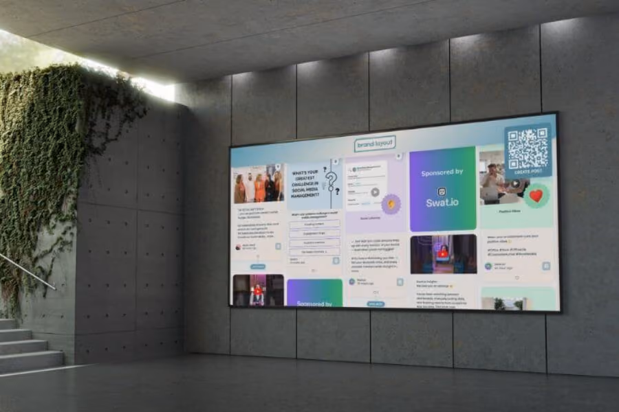 A large wall-mounted screen in a modern office displays a branded social wall with polls, sponsored content, and a QR code for creating posts.