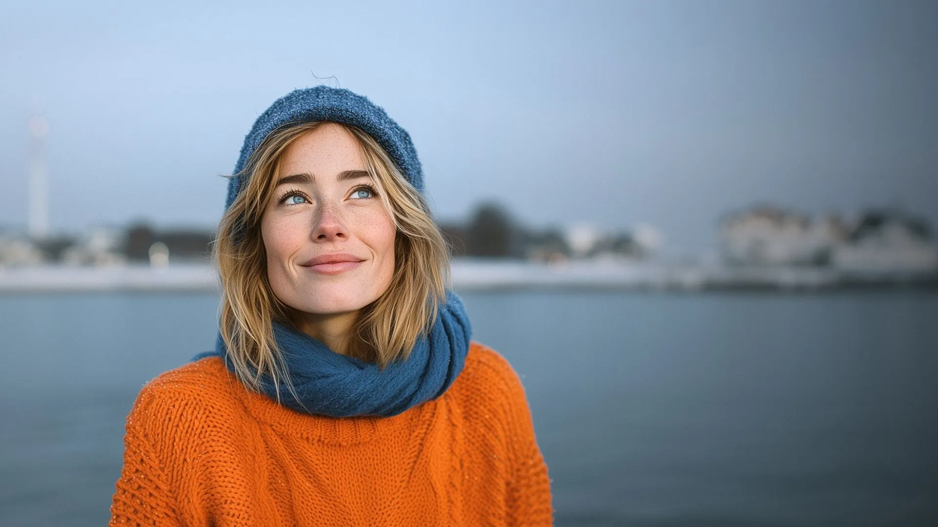 Smiling woman with blue eyes wearing a blue knit hat and scarf and an orange sweater, standing near a body of water.