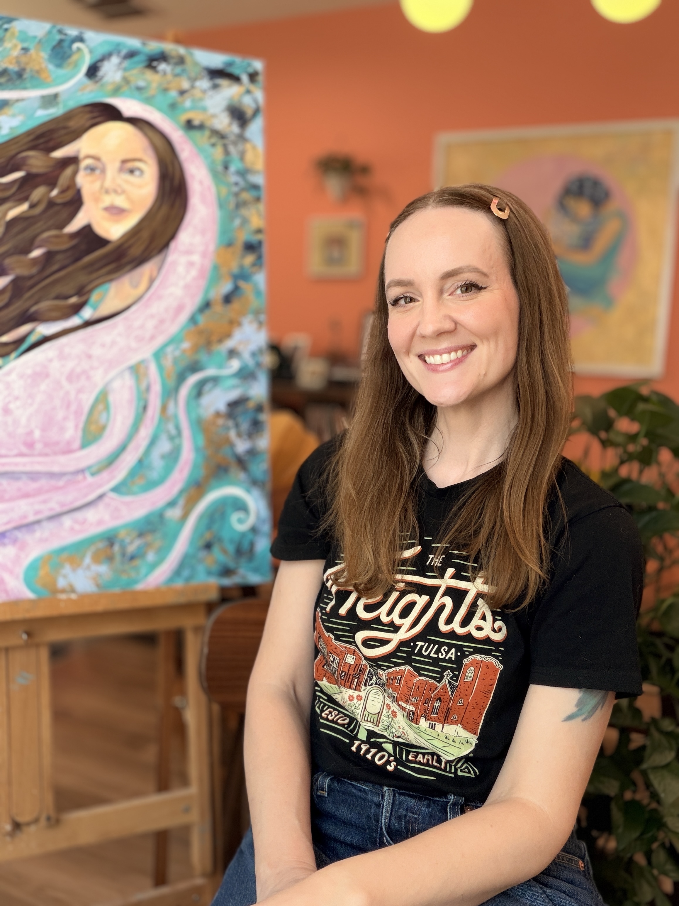 A photograph of Shayna Brewer. She is wearing a black t-shirt that says "The Heights, Tulsa" and smiling. There is a colorful background with artwork behind her that is blurred by the short focus lens.