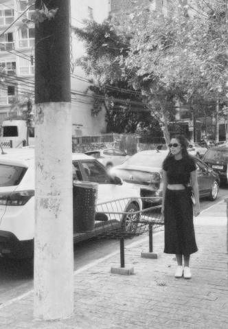 Foto minha em pé em uma calçada olhando para frente. Na rua tem carros estacionados. Estou de saia e cropped pretos, óculos de sol e tenis branco.