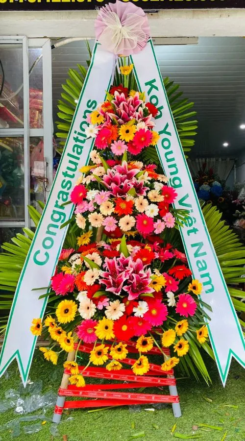 Màu sắc: đỏ tươi rực rỡ. Dịp tặng: khai trương, khánh thành, tân gia, chúc mừng thành công, kỷ niệm thành lập. Loại sản phẩm: Hoa khai trương.

Hoa Khai Trương Hàn Quốc 001 là mẫu kệ hoa khai trương mang tone màu chủ đạo đỏ tươi rực rỡ, tượng trưng cho may mắn, thành công rực rỡ và tài lộc dồi dào — lựa chọn được nhiều khách hàng tin tưởng khi cần một lẵng hoa khai trương thật ấn tượng.

Thành phần hoa chính gồm lay ơn đỏ, hồng đỏ và trạng nguyên đỏ, phối cùng dương xỉ, lá bạc và baby trắng để tôn lên tổng thể tươi tắn, hài hòa và tràn đầy sức sống cho không gian khai trương.

Sản phẩm được thiết kế theo dáng kệ hoa đứng 2 tầng cân đối, form dáng vững chãi, cân đối, phù hợp trưng bày ở những vị trí trang trọng, dễ gây ấn tượng ngay từ cái nhìn đầu tiên.

Chiều cao kệ hoa khoảng 1m7 – 1m9 tùy mẫu, đường kính chân đế 55 – 65cm, kích thước lý tưởng để đặt trước sảnh, cửa ra vào hoặc khu vực sự kiện mà không chiếm quá nhiều diện tích.

Đây là món quà thích hợp gửi tặng trong các dịp khai trương cửa hàng, công ty, spa, khách sạn, nhà hàng, văn phòng hay showroom, giúp chủ nhân buổi lễ thêm phần rạng rỡ và trang trọng.

Với thiết kế sang trọng, Hoa Khai Trương Hàn Quốc 001 là mẫu kệ hoa khai trương, lẵng hoa khai trương được yêu thích trong nhóm hoa khai trương sang trọng, hoa khai trương cao cấp dành cho khách hàng doanh nghiệp.

Sản phẩm gửi gắm thông điệp chúc mừng ý nghĩa: chúc công việc kinh doanh thuận buồm xuôi gió, làm ăn phát đạt, vạn sự hanh thông và ngày càng thịnh vượng. Dải băng rôn có thể tùy chỉnh nội dung lời chúc theo yêu cầu.

Liên hệ Shop Hoa Mãi Mãi ngay hôm nay để được tư vấn chọn mẫu phù hợp và đặt hoa giao nhanh tận nơi!