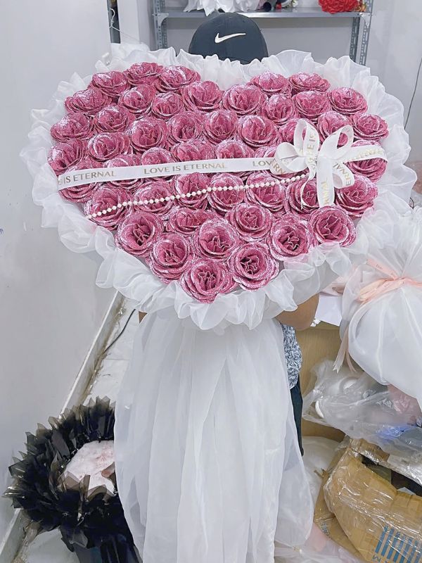 Màu sắc: hồng tím khói ánh nhũ. Dịp tặng: Valentine, kỷ niệm, tỏ tình, tặng vợ. Loại sản phẩm: Hoa sáp.

Điểm riêng của Hoa Sáp Thơm Tình Yêu nằm ở mùi hương: từng bông sáp được ướp thơm dịu, để trong phòng kín vẫn thoảng mùi sau nhiều tuần. Về màu, bó đi theo sắc hồng tím khói trầm, cánh phủ nhũ nên bắt sáng rất đẹp mà không bị chói.

Hoa được kết thành khối trái tim gồm khoảng 55 đến 65 bông, cánh xoắn tròn nhiều lớp, mặt tim đầy và bo viền gọn.

Quanh viền là voan trắng bèo nhún với đuôi voan buông dài phía dưới, mặt hoa vắt một dây ngọc trai trắng và dải ruy băng in chữ "Love Is Eternal". Trái tim rộng khoảng 40 đến 45cm, cao chừng 55cm tính cả phần voan, ôm vừa một vòng tay.

Mẫu này hợp tặng vợ, người yêu hoặc bạn gái vào dịp Valentine, kỷ niệm ngày cưới, ngày quen nhau, nhất là với những người thích quà có mùi hương riêng.

Anh chị đặt bằng cách gọi 0934.833.013 – 0907.473.357 hoặc nhắn Facebook Hoa Mãi Mãi, shop cho chọn hương và đổi tông màu hoa theo ý.