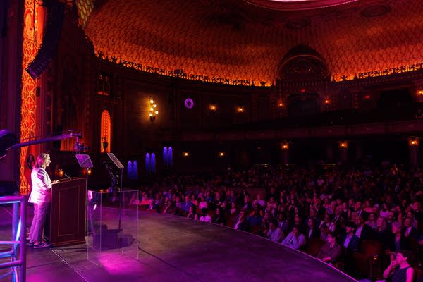 Wide shot of a woman standing on stage at a podium, looking out at a theater full of attendees.