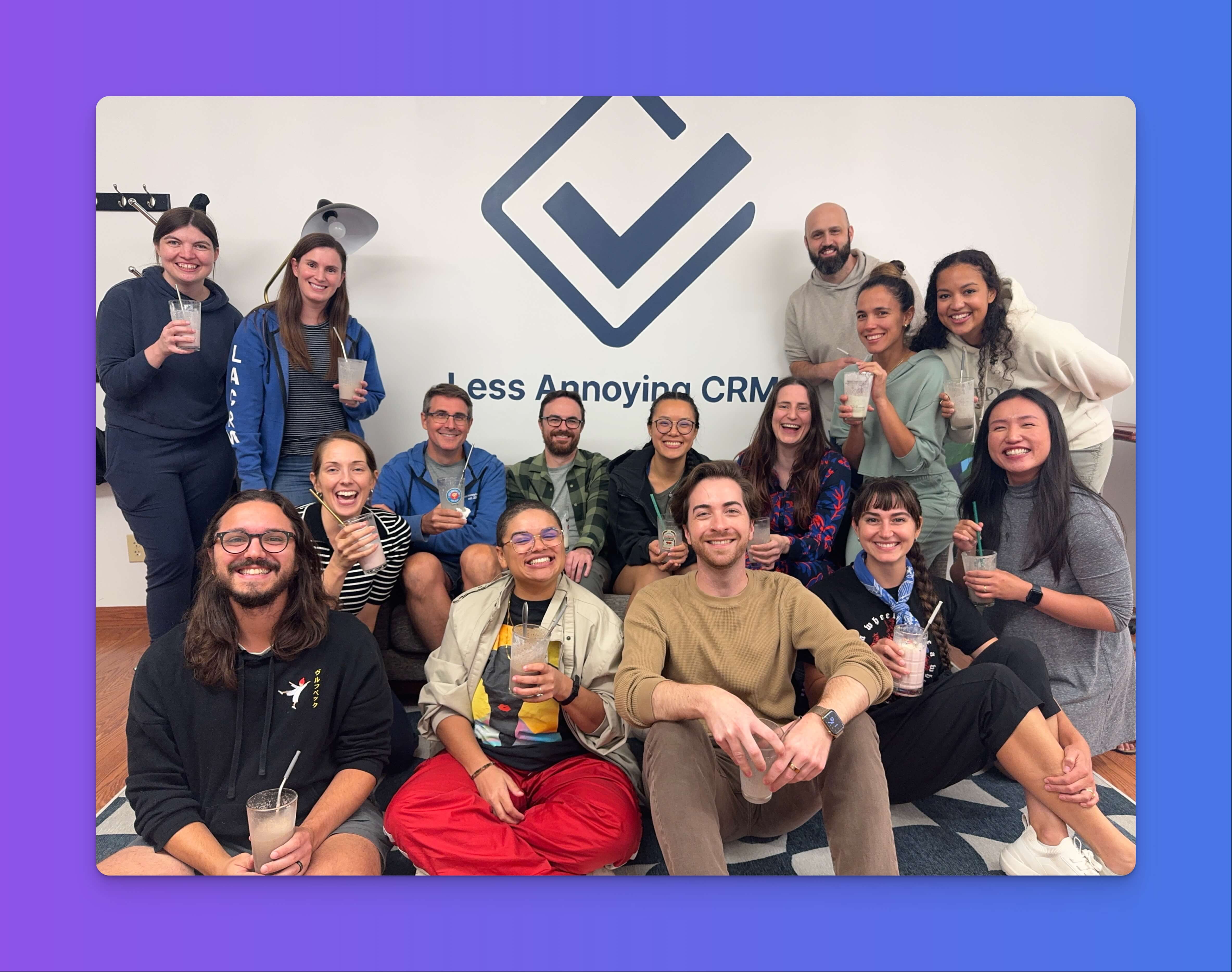 The Less Annoying CRM team smiling and holding up milkshakes in front of the company logo on the office wall