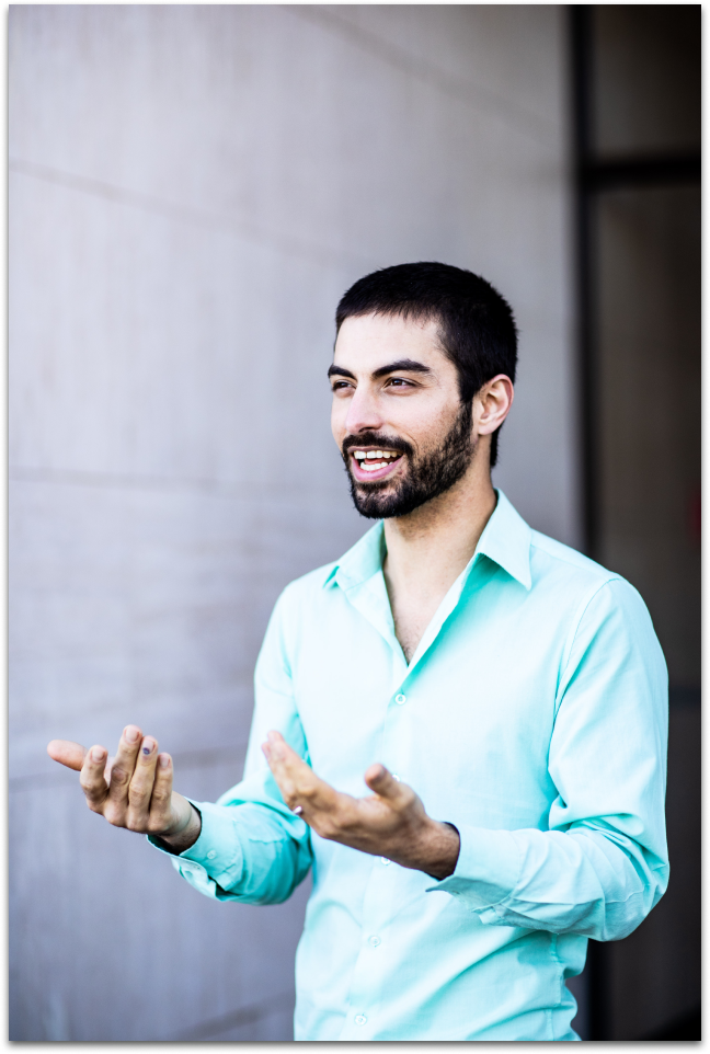 Smiling dark-haired bearded man in a teal collared shirt with hands in front, palms upward as in conversation
