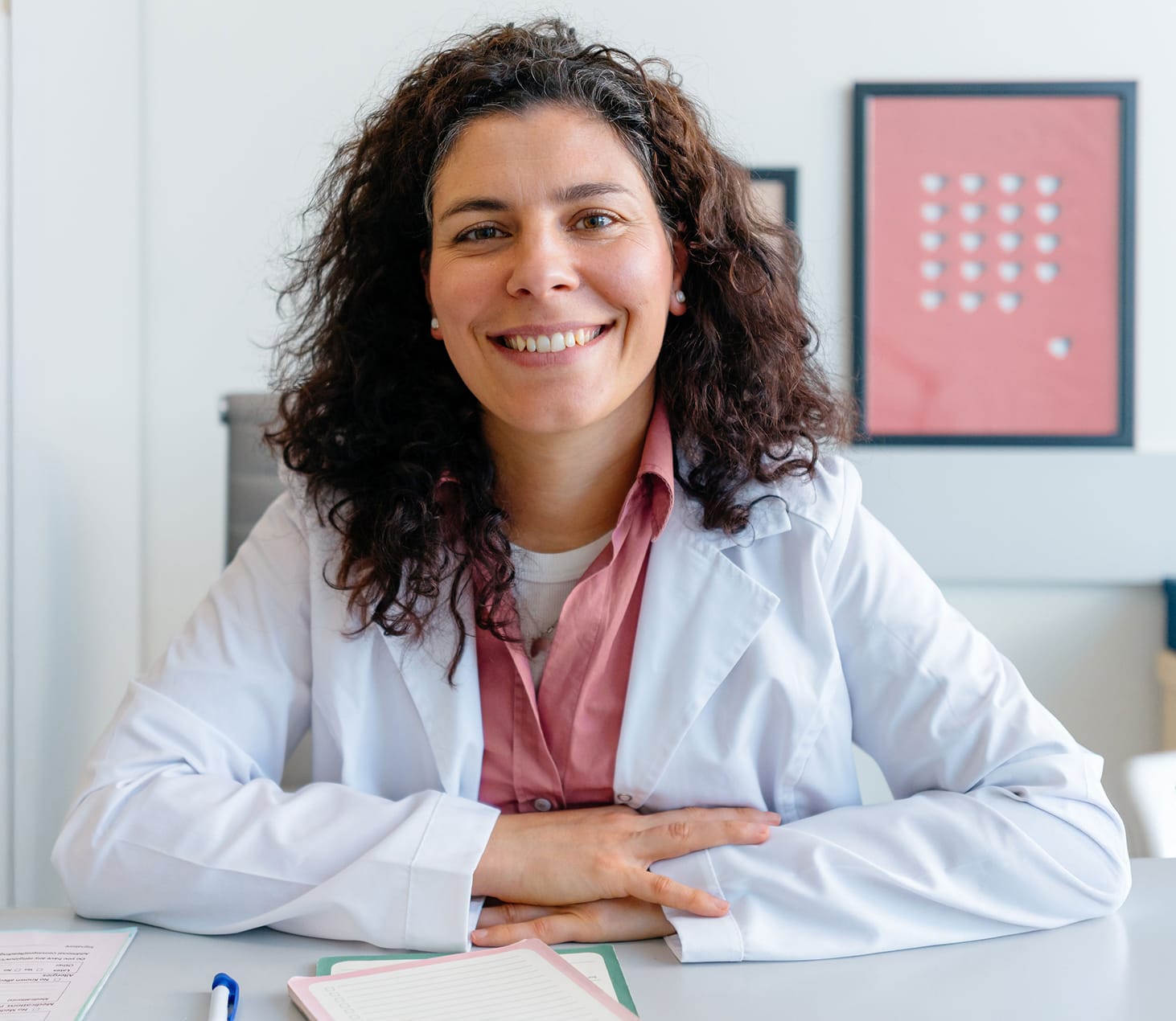 Smiling female doctor with curly hair wearing a white lab coat sitting at a desk with papers and a pen.