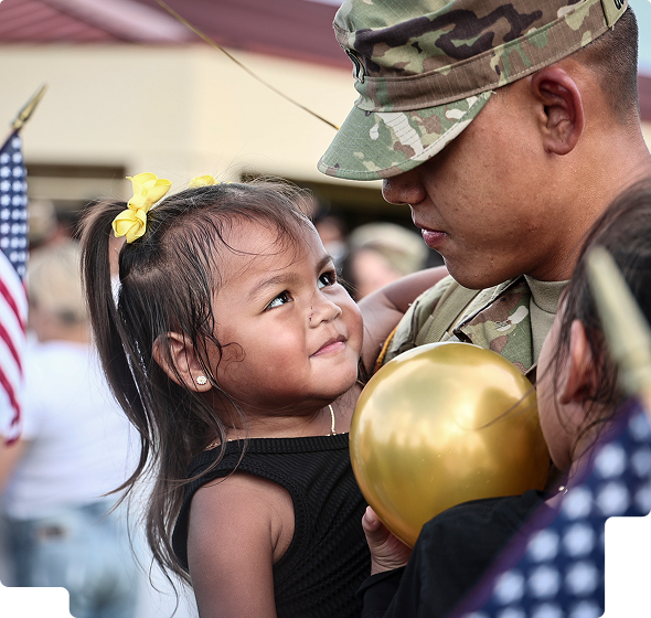 Young girl with yellow hair bows gazes affectionately at a soldier in camouflage uniform holding a gold balloon.