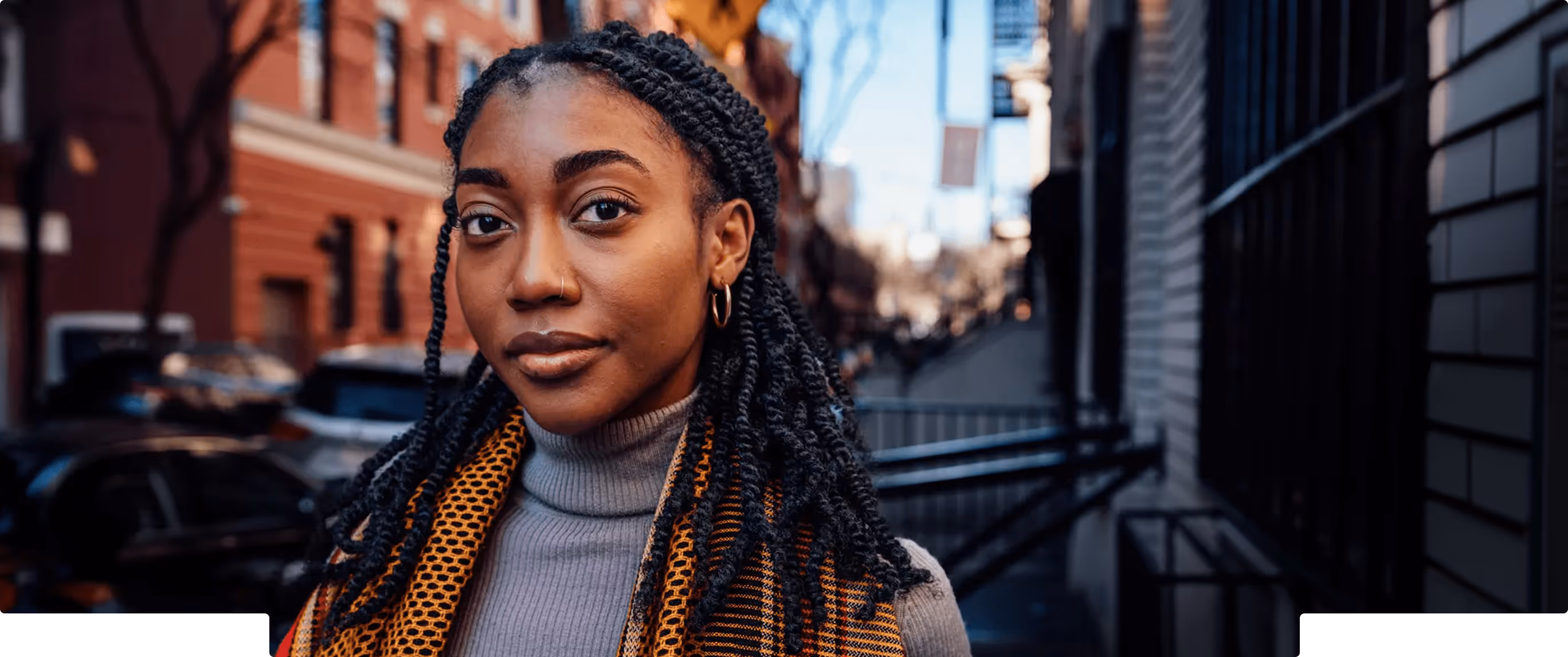 Close-up of a young woman with braided hair and hoop earrings wearing a gray turtleneck and patterned scarf, standing on a city street.