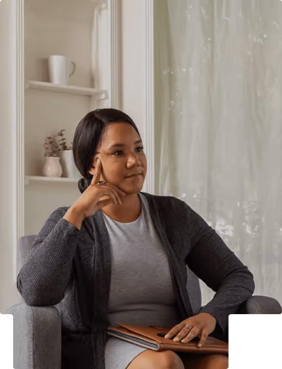 Thoughtful woman seated in an armchair holding a notebook, with a cozy interior background.