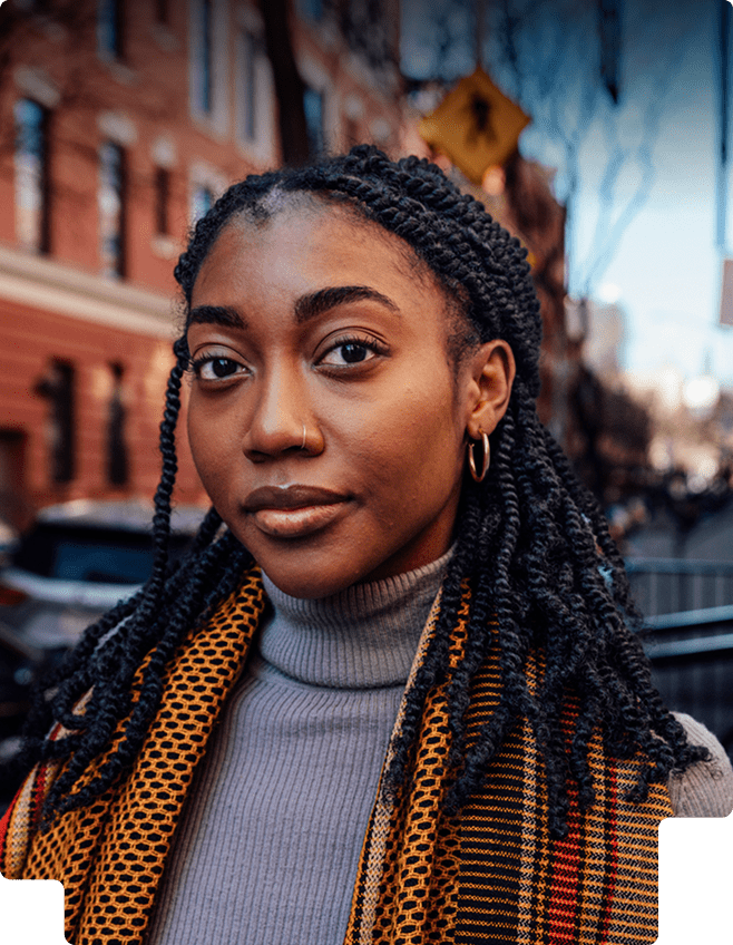 Close-up portrait of a woman with long braided hair, wearing gold hoop earrings, a gray turtleneck, and a patterned scarf outdoors in an urban setting.