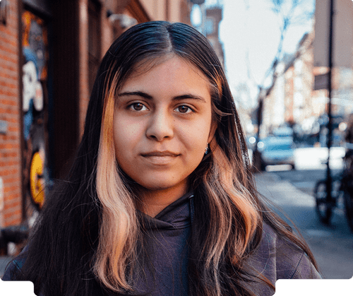 Photo of a young girl wearing a sweatshirt slightly smiling at the camera in an urban setting.