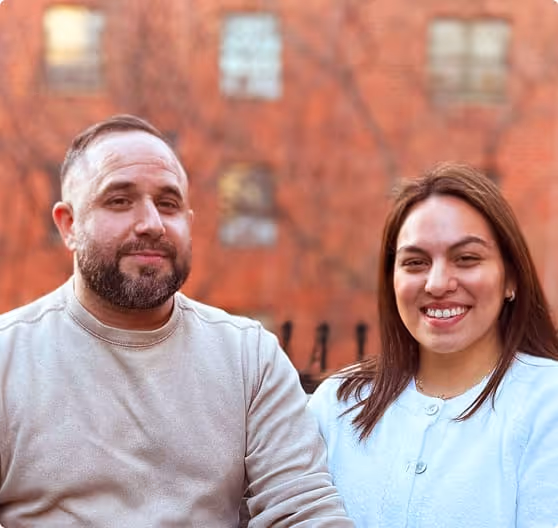 Smiling couple sitting outdoors with a brick building in the background.