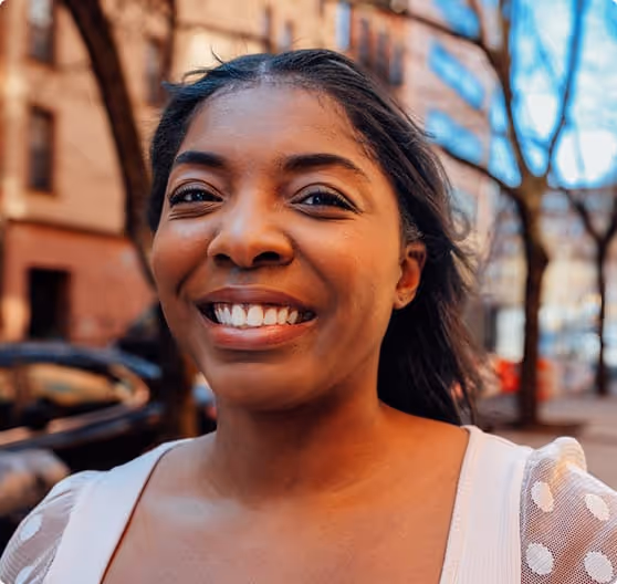 Smiling young woman with dark hair standing outdoors on a city street with buildings and trees in the background.