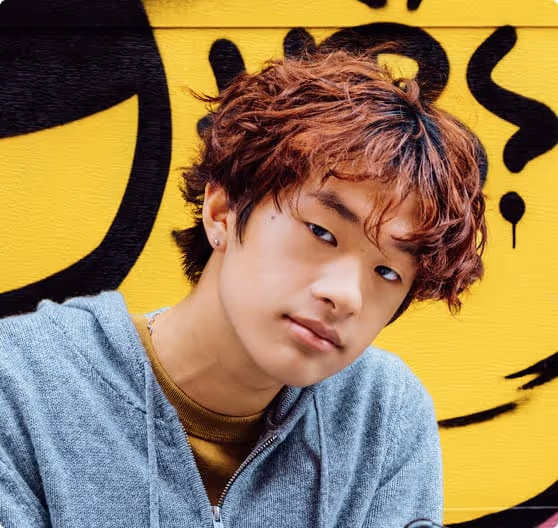 Young man with curly reddish-brown hair wearing a gray hoodie, posing in front of a yellow wall with black graffiti.