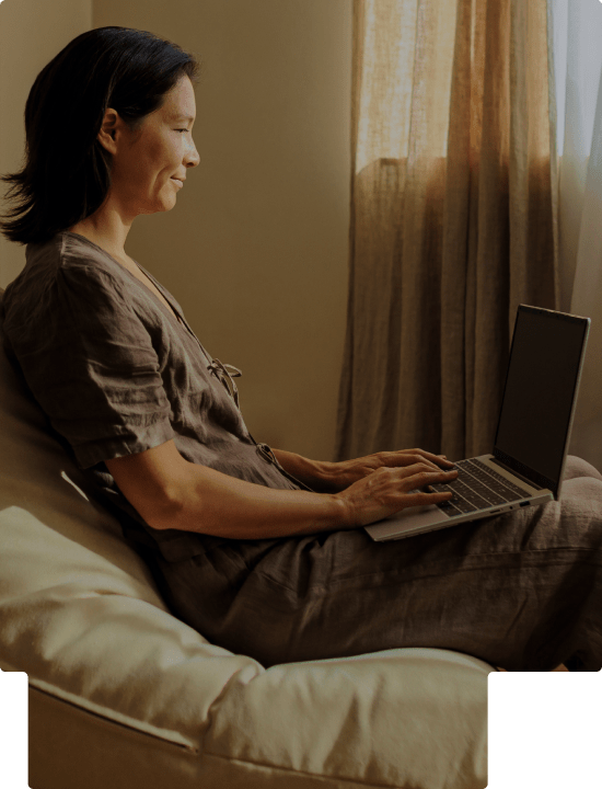 Woman sitting on a cushioned chair typing on a laptop with soft natural light from a window.