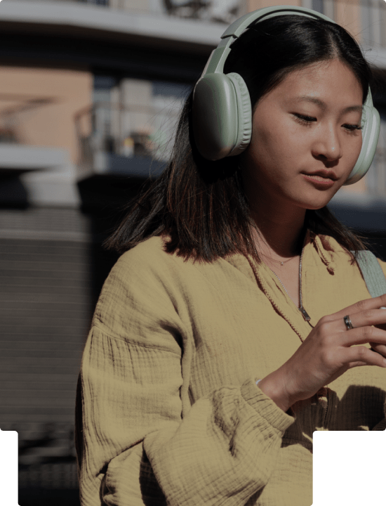 Young woman wearing green headphones and a beige jacket, focused on a smartphone in her hands outdoors.