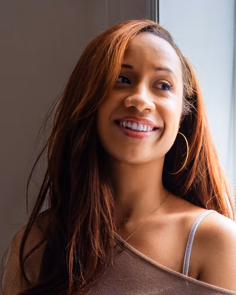 Smiling woman with long auburn hair, wearing a one-shoulder top, hoop earrings, and a nose ring, looking to the side near a window.
