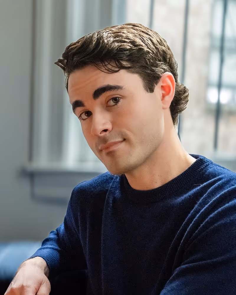 Young man with brown hair wearing a navy sweater sitting indoors near a window.