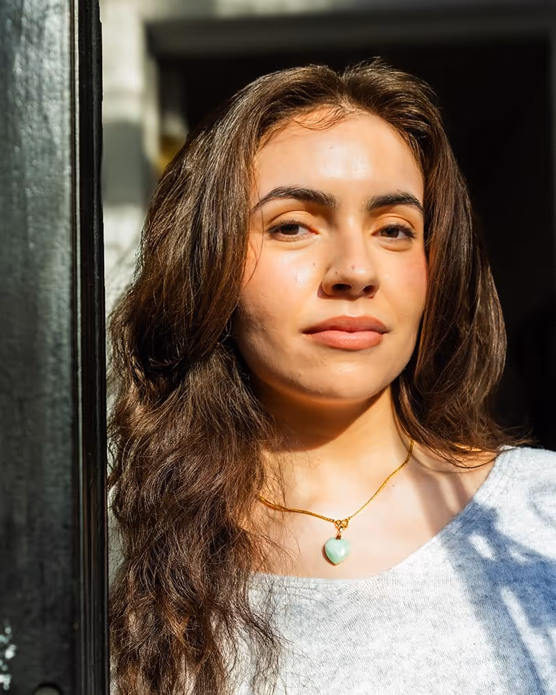 Woman with long brown hair wearing a gray top and a gold necklace with a heart-shaped pendant, illuminated by sunlight.