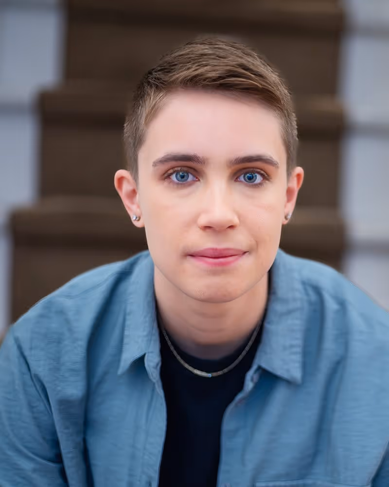 Portrait of a person with short light brown hair and blue eyes wearing a blue shirt and a small chain necklace.