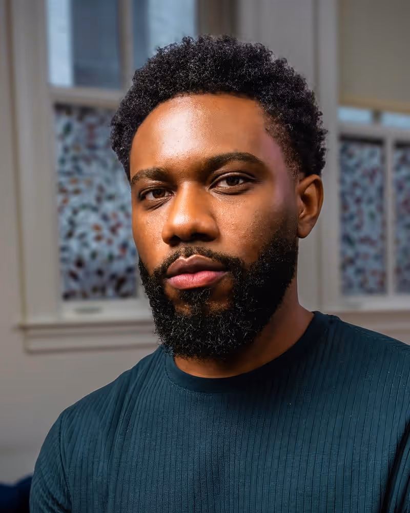 Close-up portrait of a young man with a full beard and curly hair wearing a dark ribbed shirt indoors.