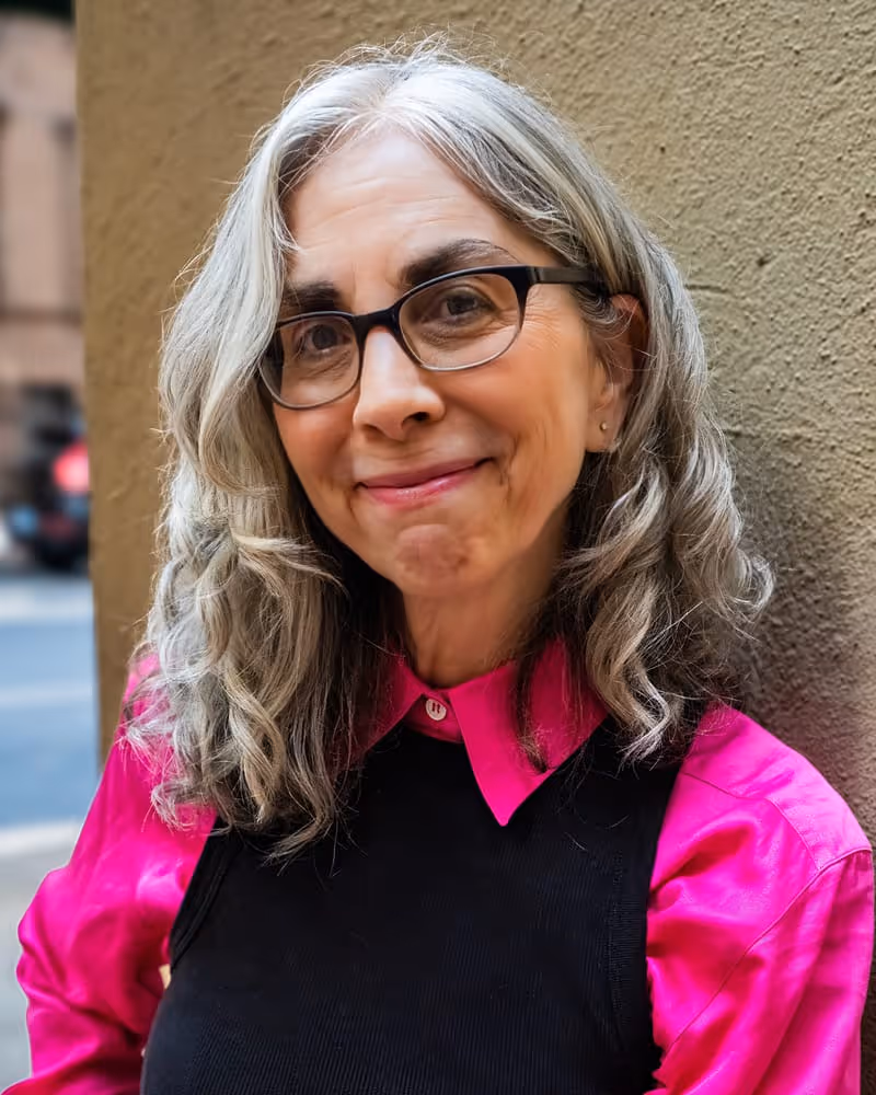 Smiling woman with gray wavy hair, wearing glasses, a bright pink shirt, and a black vest standing outdoors next to a textured wall.
