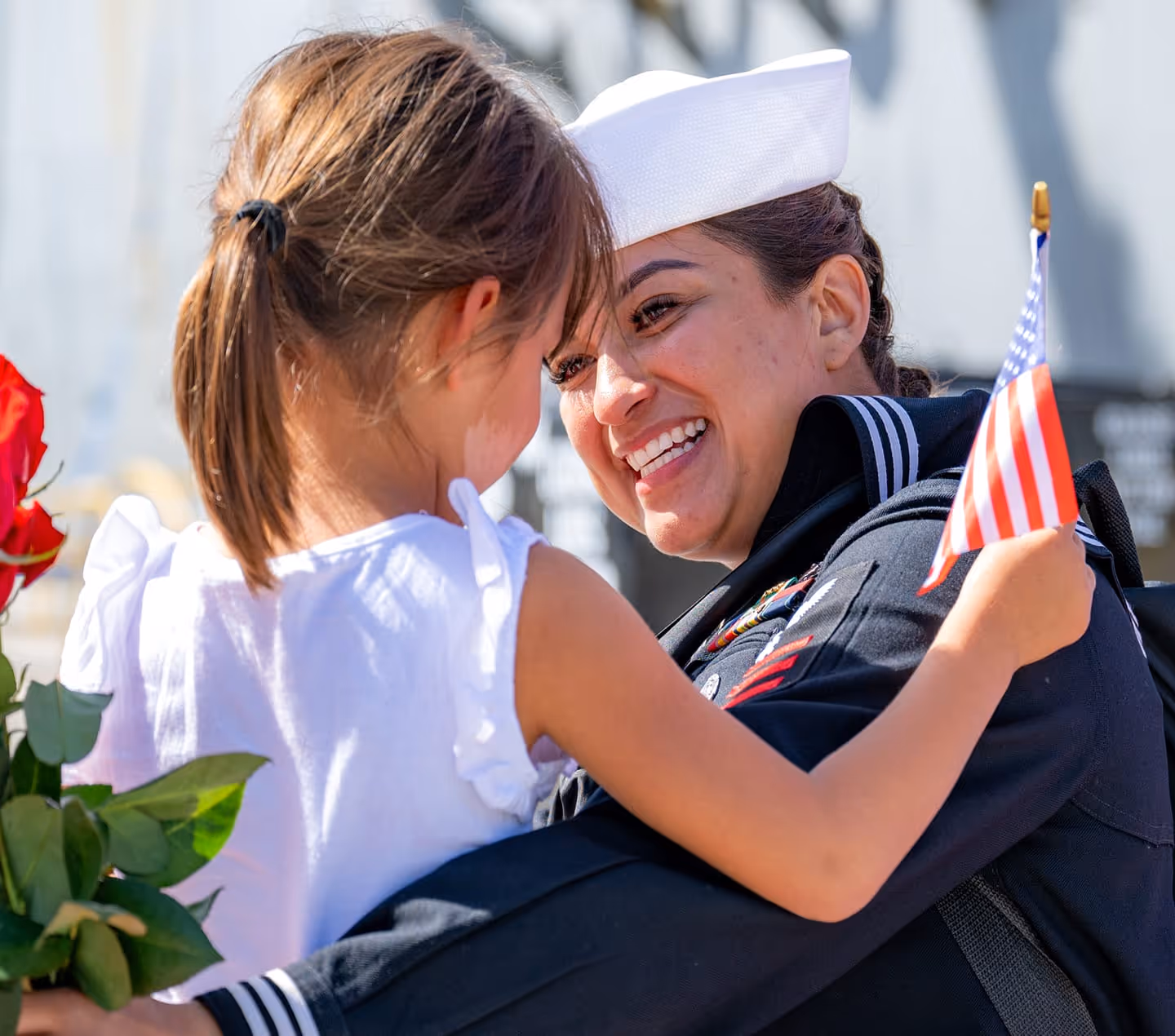 Smiling female sailor in uniform embracing a young girl holding an American flag and a red rose.