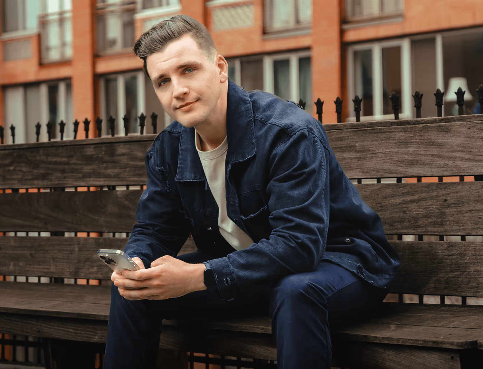 Man sitting on wooden bench outdoors, holding a smartphone and looking at the camera.