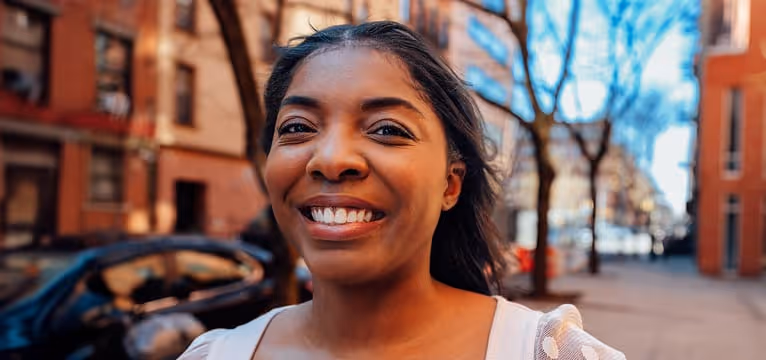 Smiling young woman outdoors with buildings and bare trees in the blurred background.