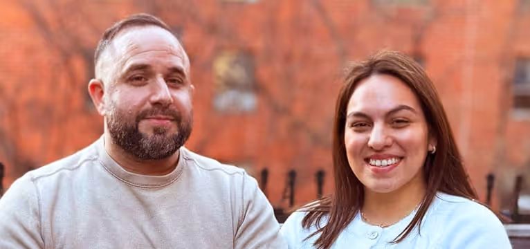Smiling man and woman sitting outdoors with blurred autumn trees and brick wall background.