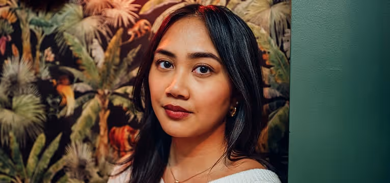 Portrait of a woman with long dark hair, wearing gold hoop earrings and a white top, against a tropical background.