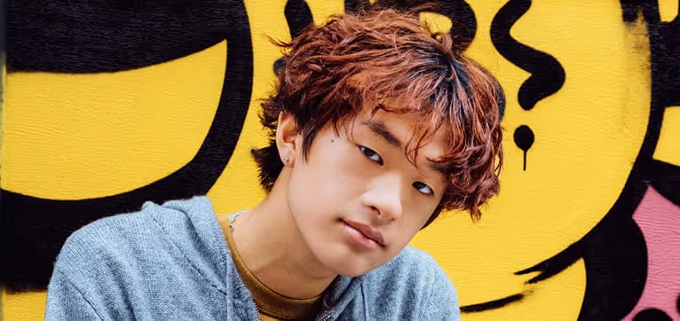 Teenage boy with curly auburn hair wearing a gray hoodie in front of a yellow graffiti wall.
