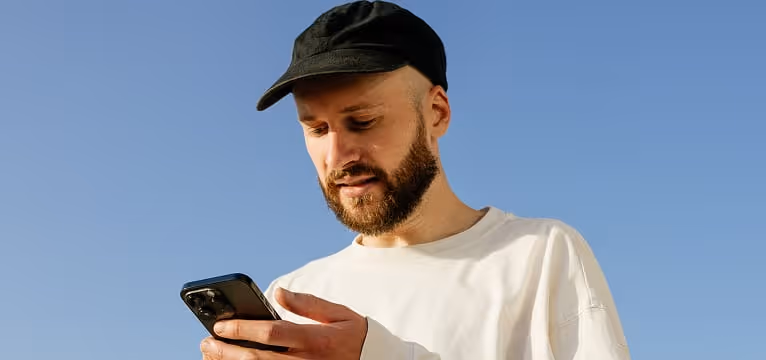 Bearded man wearing black cap and white shirt looking at his smartphone against clear blue sky.