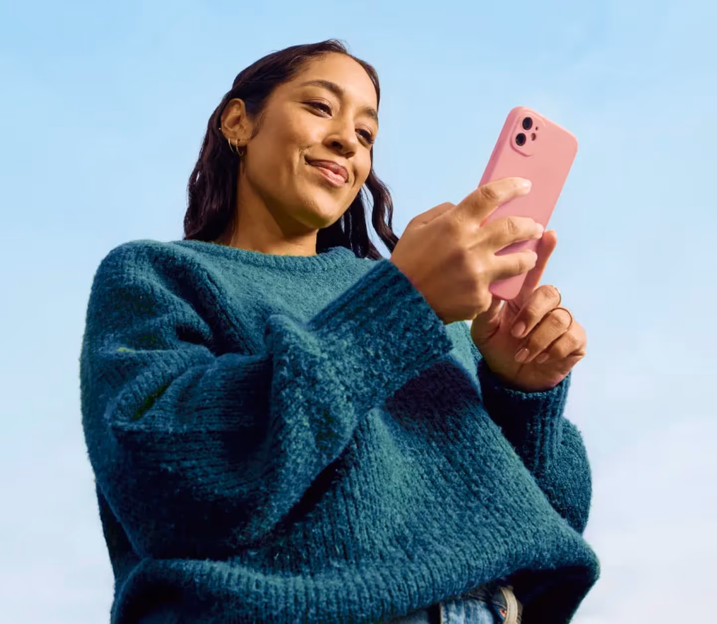 Woman in teal sweater smiling while holding and looking at a pink smartphone against blue sky.