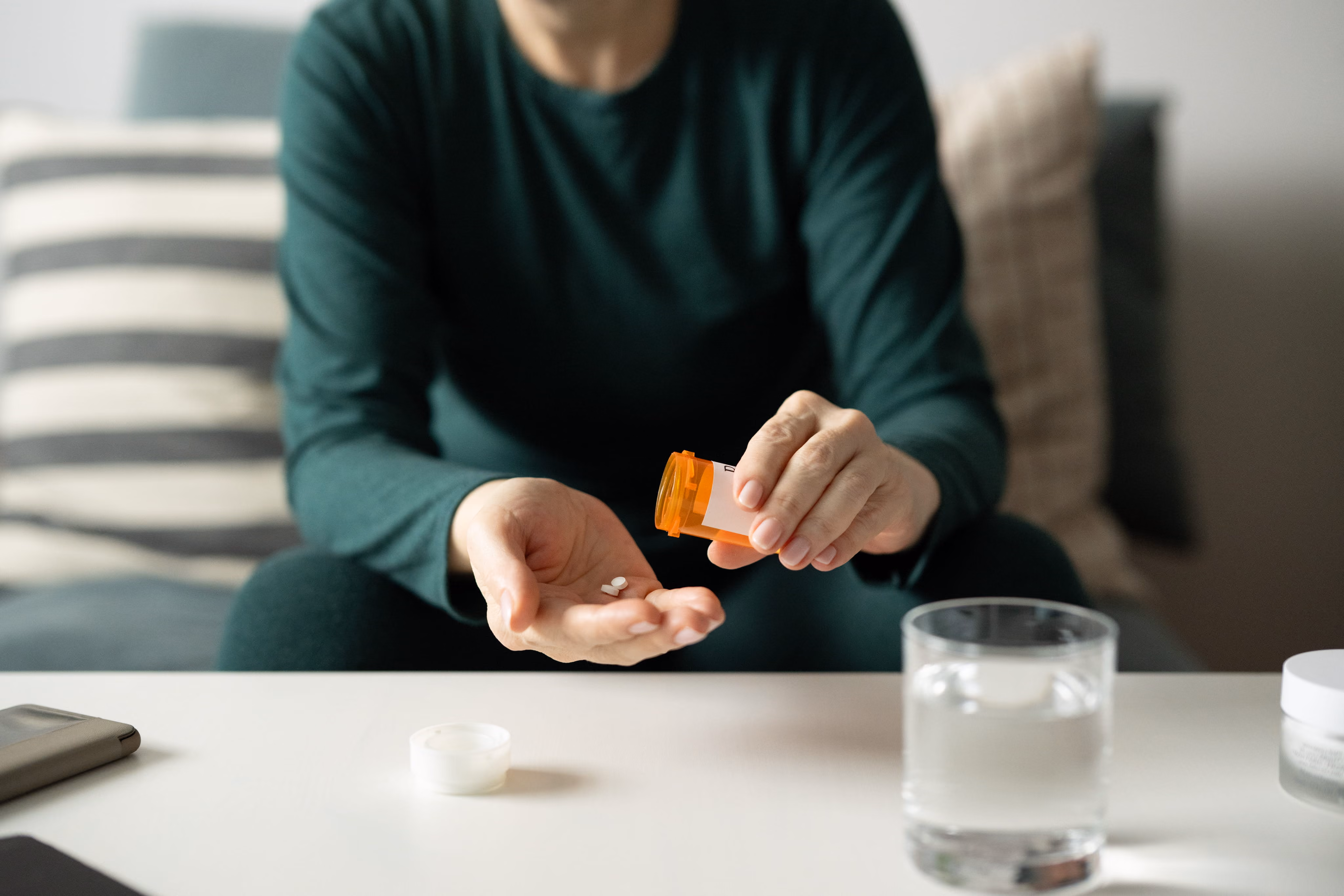 Person in green shirt pouring white pills from an orange prescription bottle into their hand with water nearby.