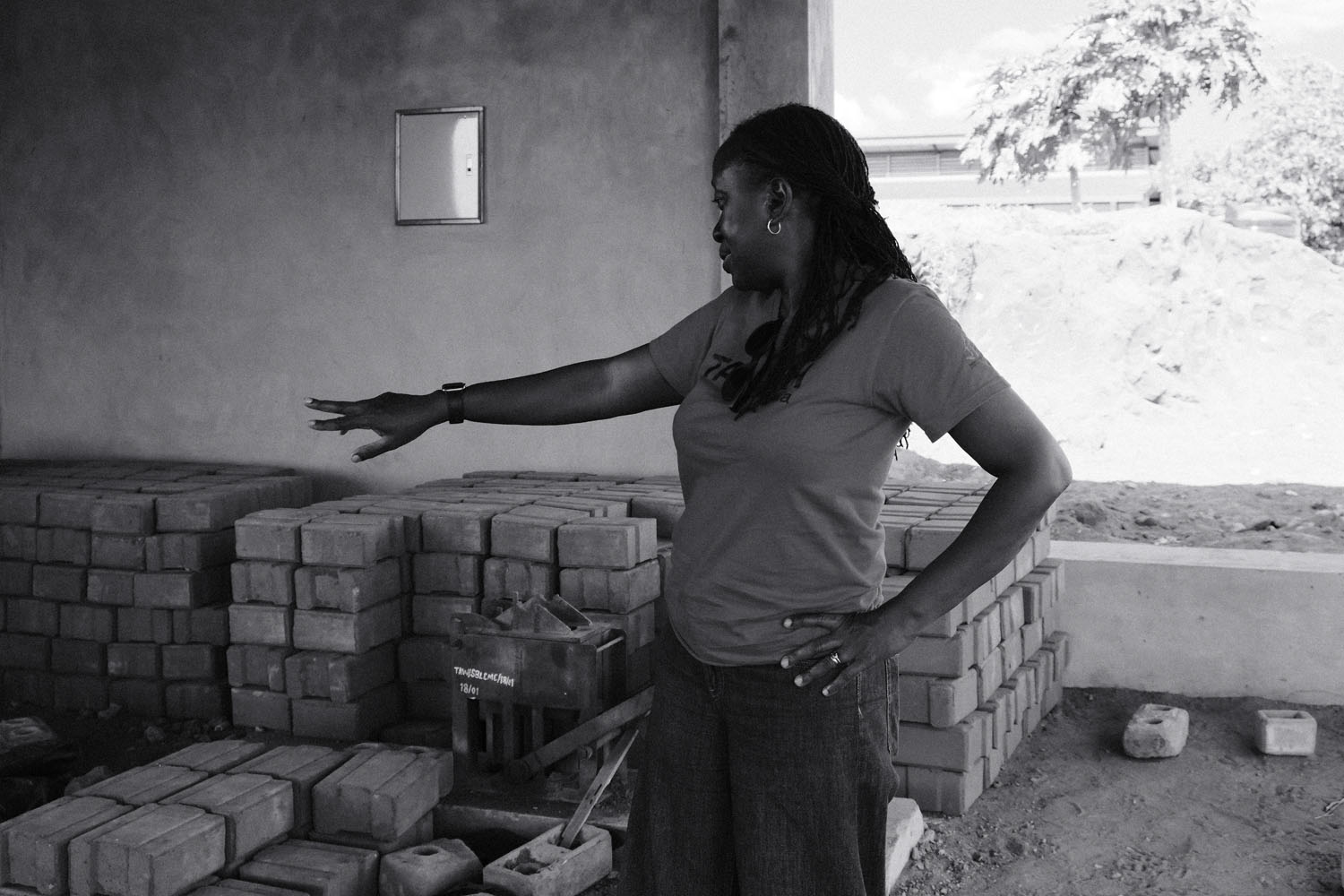 Victoria gesturing towards a hand-operated brick press surrounded by stacked bricks