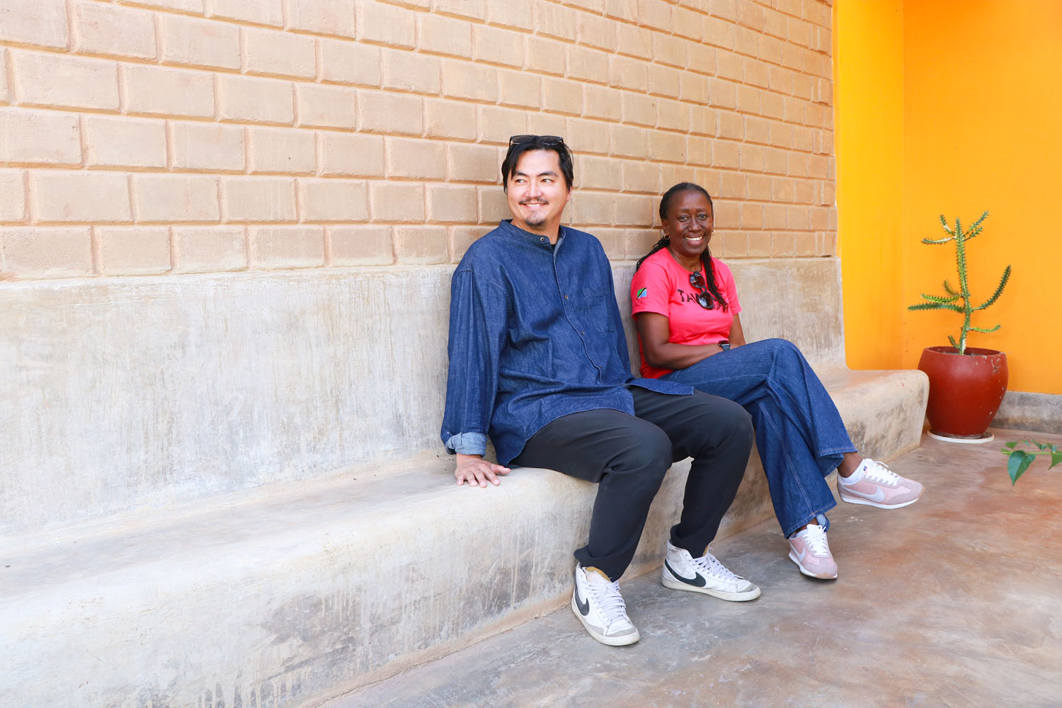 Jun and Victoria Marwa Heilman sitting on a concrete bench against an earth-brick wall