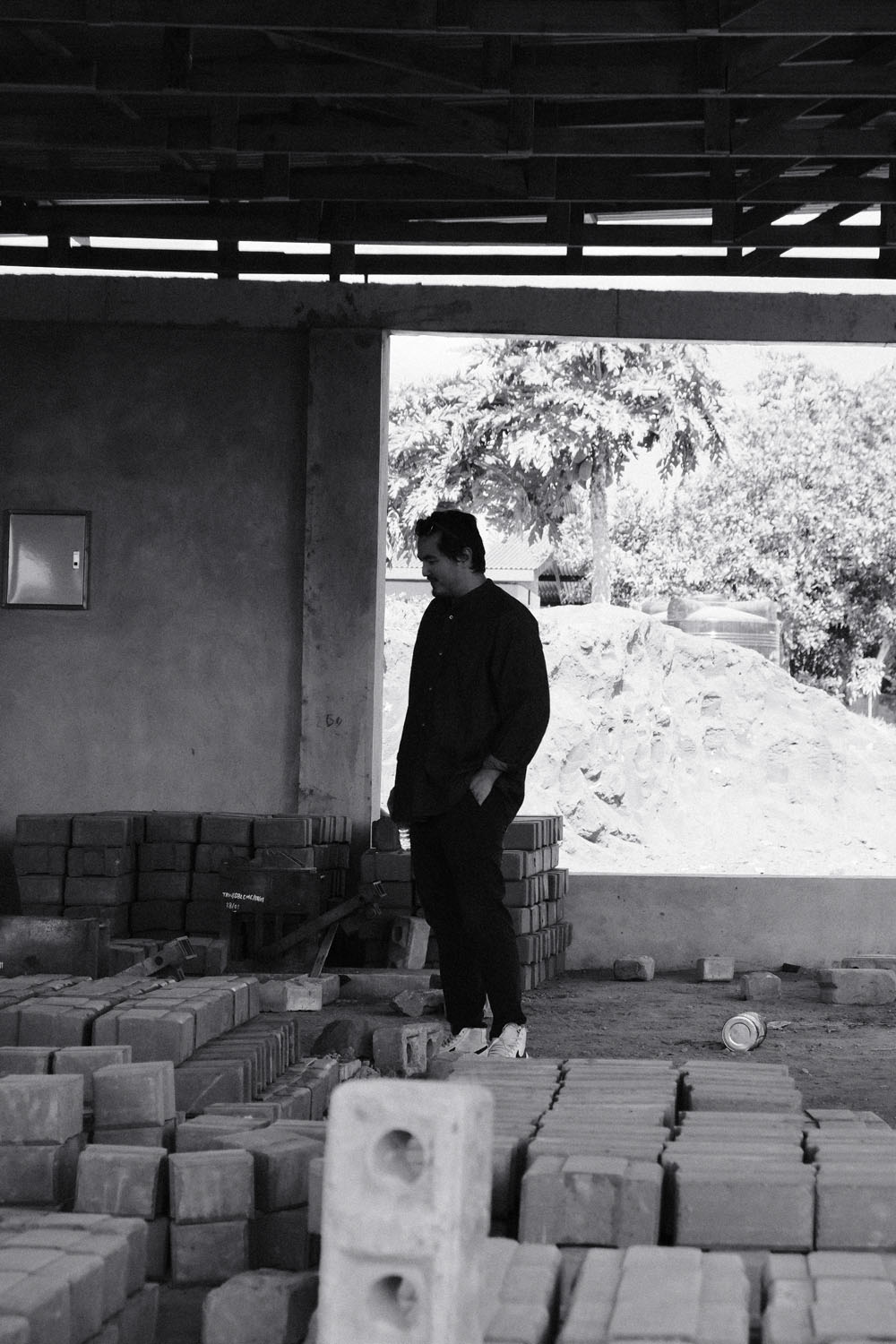 Jun standing among stacks of earth bricks, looking down at them