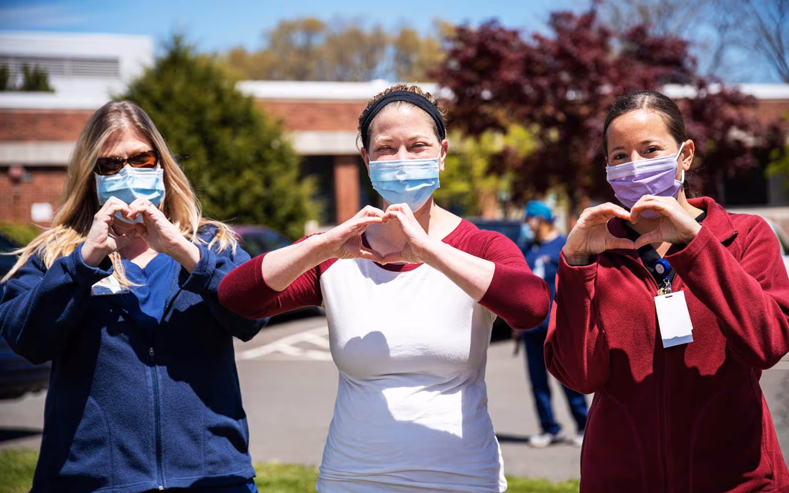 Three women wearing masks making hearts with their hands.