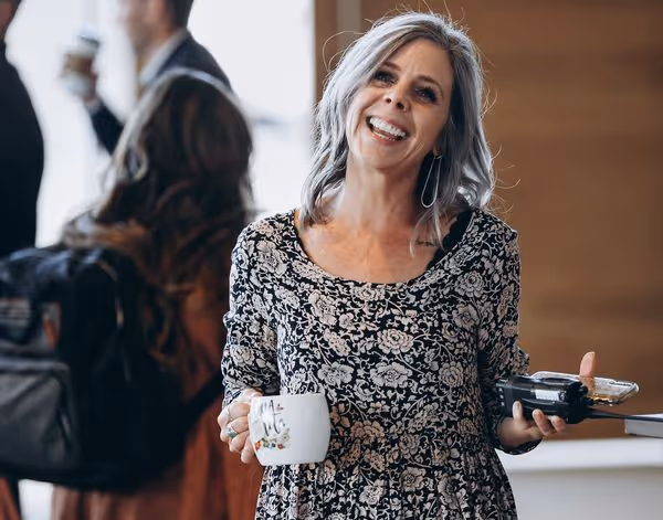 A woman smiles and tilts her head with a coffee mug in her hand